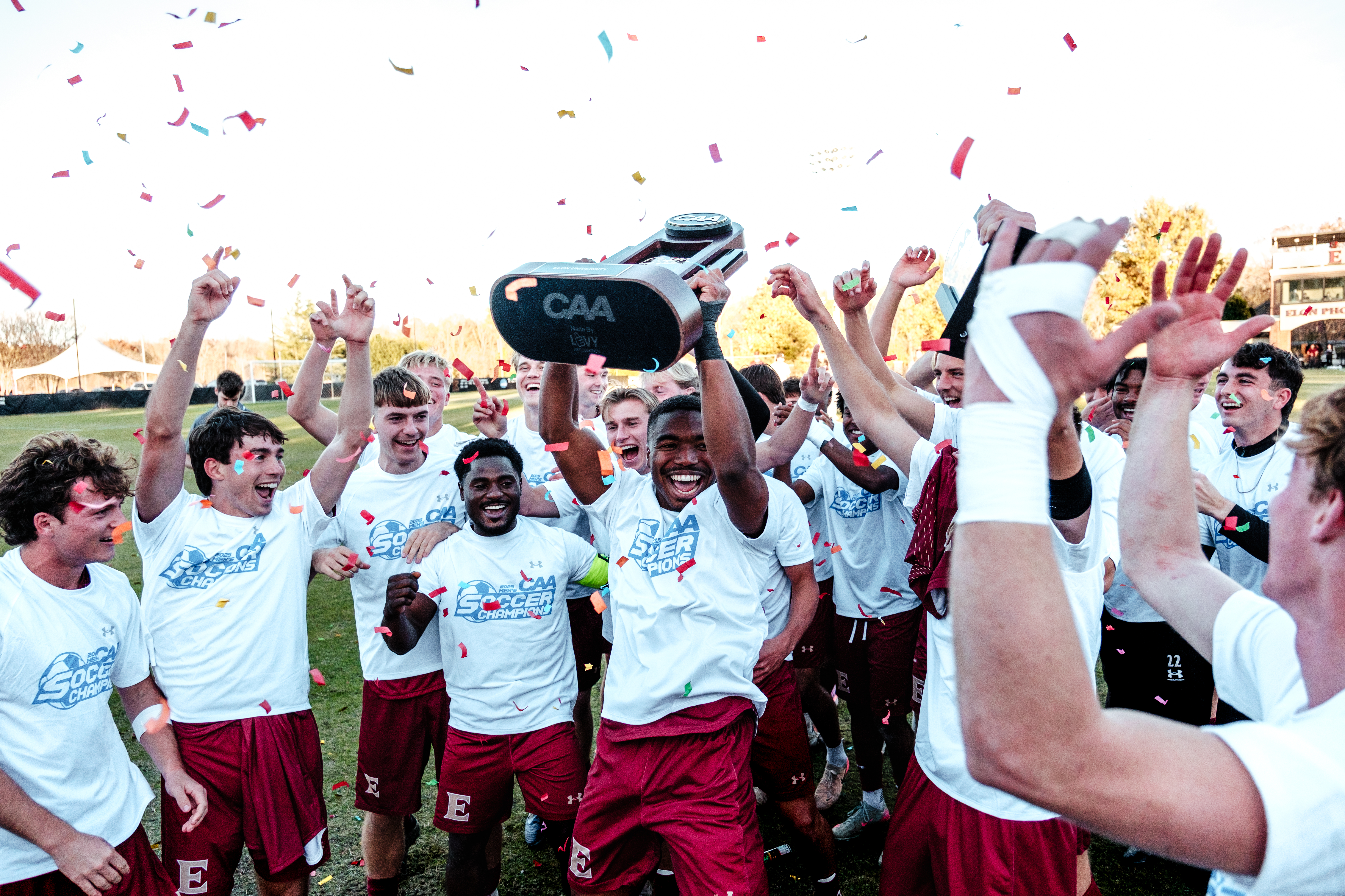 Elon soccer players celebrate a win while cheering together. One player in the middle holds up a trophy