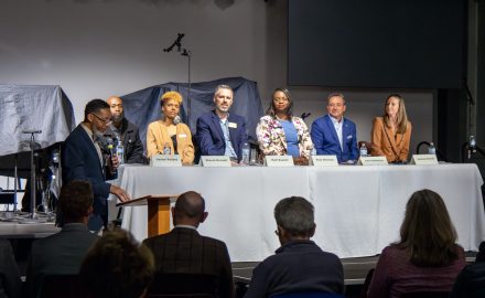 A man speaks at a podium in front of a well-lit table of six panelists. The backs of audience members' heads are in the foreground