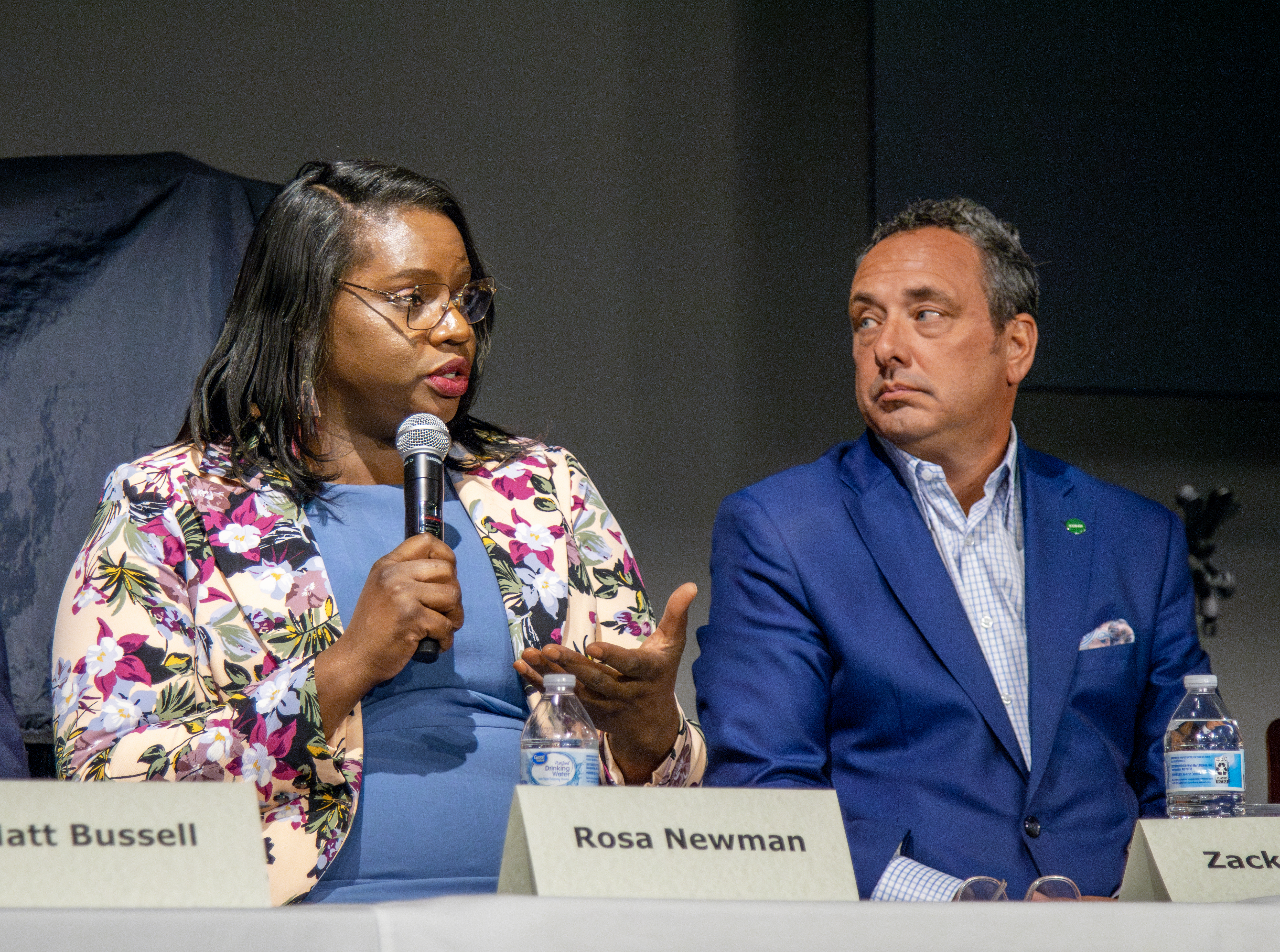 A woman in a floral blazer and blue blouse speaks into a microphone as a man in a blue suit is turned toward her listening.