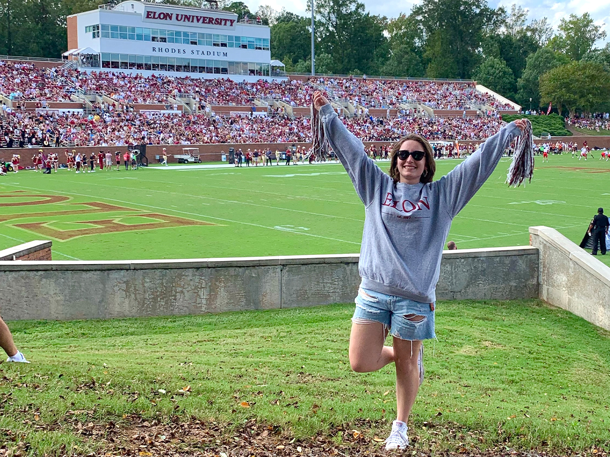 A girl has her hands raised in front of a football field while wearing an Elon sweatshirt