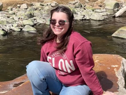 Woman in sunglasses and Elon shirt sits on a rock in a river