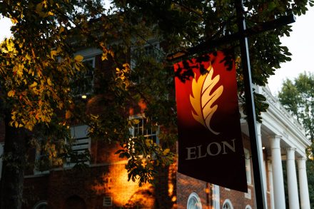 A red Elon University banner with a golden oak leaf logo hangs among autumn branches, illuminated by warm evening light in front of a brick campus building with white columns.