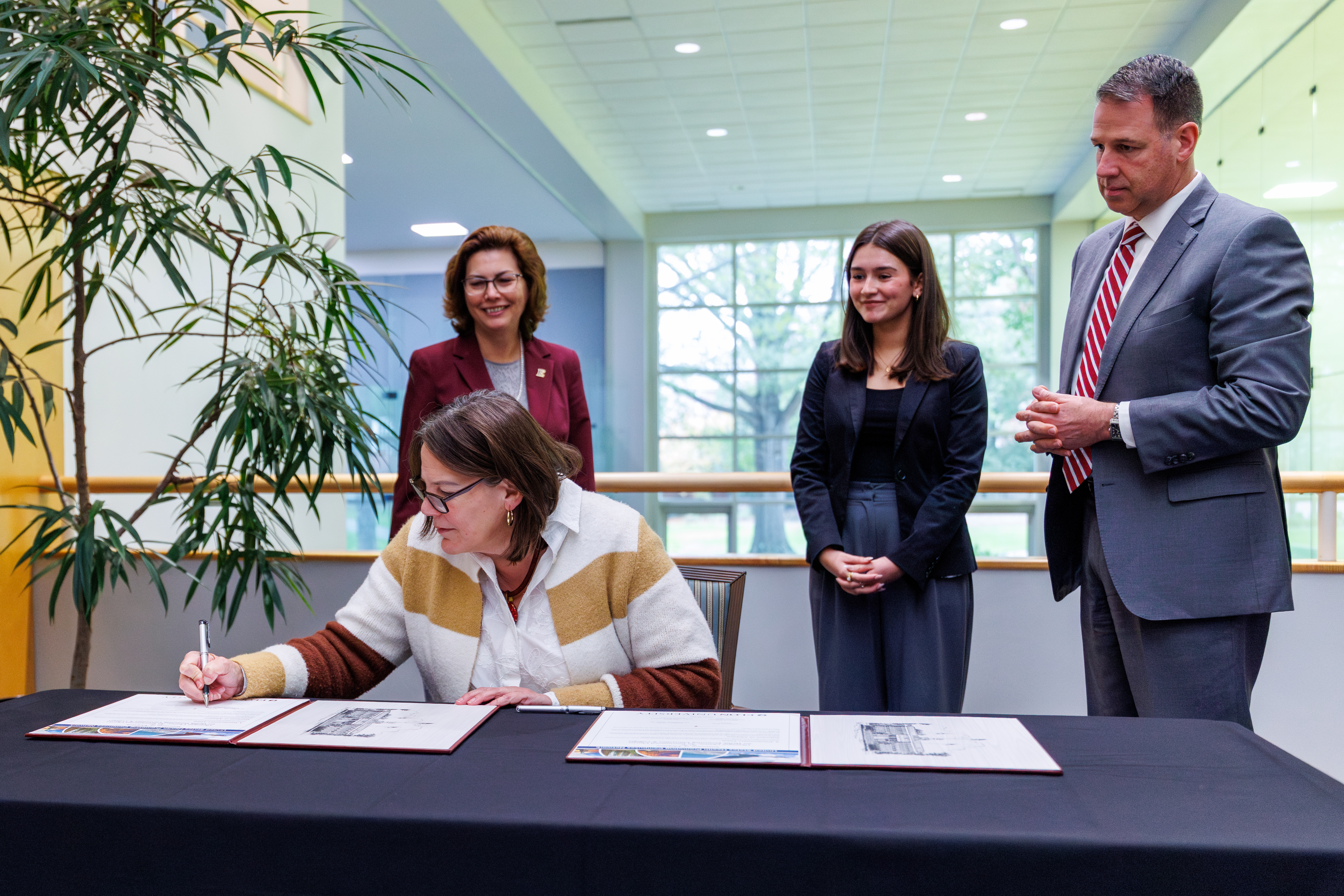 A university leader signs a framed document on a table during a formal ceremony as three others stand nearby, smiling and observing. The event takes place in a bright indoor space with large windows and greenery in the background.
