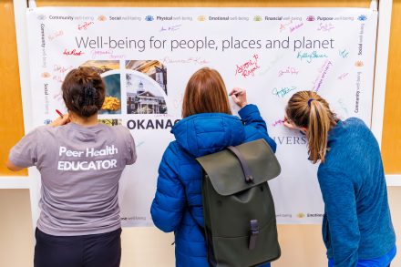 Three people sign a large banner that reads “Well-being for people, places and planet” during an event at Elon University. One person wears a “Peer Health Educator” shirt, while others in jackets and backpacks add their signatures in colorful ink around the Okanagan Charter display.