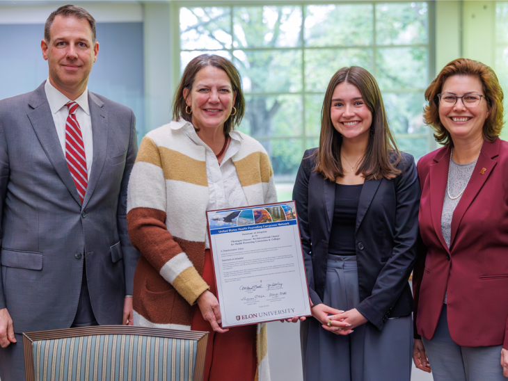 Four people stand together smiling as one holds a framed document from Elon University. The document marks the university’s adoption of the Okanagan Charter for health-promoting campuses. The group stands in a bright room with large windows and greenery visible outside