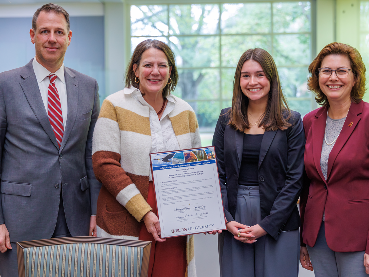 Four people stand together smiling as one holds a framed document from Elon University. The document marks the university’s adoption of the Okanagan Charter for health-promoting campuses. The group stands in a bright room with large windows and greenery visible outside