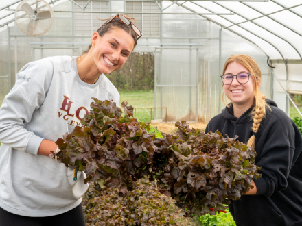 Two smiling students hold large bundles of freshly harvested lettuce inside a greenhouse. One wears an Elon volleyball sweatshirt, and the other a black hoodie with a braid. Rows of leafy greens and greenhouse panels fill the background.