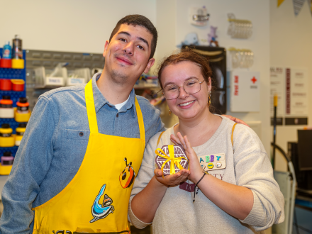 Two students smile in the Maker Hub, one wearing a yellow apron decorated with colorful patches and the other holding a handmade project tied with a yellow ribbon. Shelves of supplies and craft materials fill the background, creating a bright and creative atmosphere.