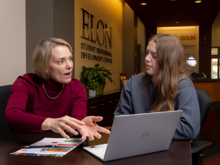 A career advisor speaks with a student inside the Elon Student Professional Development Center. The advisor gestures while explaining something, and the student listens attentively with an open laptop and documents on the table in front of them.