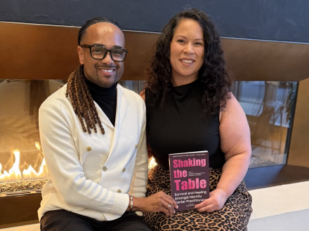 Two people sit together in front of a modern fireplace, smiling and holding a book titled “Shaking the Table: Survival and Healing Amongst Identity Center Practitioners.” The setting appears warm and professional, with soft lighting and contemporary decor.