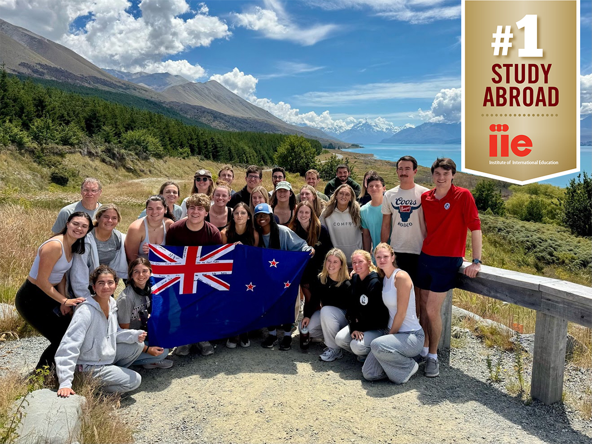 A group of people hold a New Zealand flag in front of mountains. A banner is on the photo in the top right corner that says 