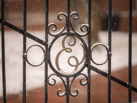 Close-up of an ornate black iron gate with the letter “E” at its center, lightly dusted with snow. The blurred background shows a brick walkway covered in snowflakes falling gently.