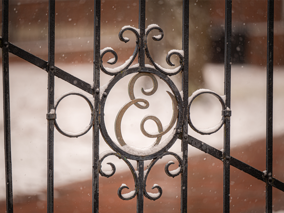 Close-up of an ornate black iron gate with the letter “E” at its center, lightly dusted with snow. The blurred background shows a brick walkway covered in snowflakes falling gently.