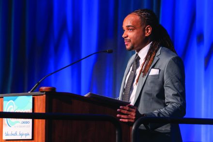 Jonathan McElderry has long hair and wears a suit and tie as he stands in front of a podium.