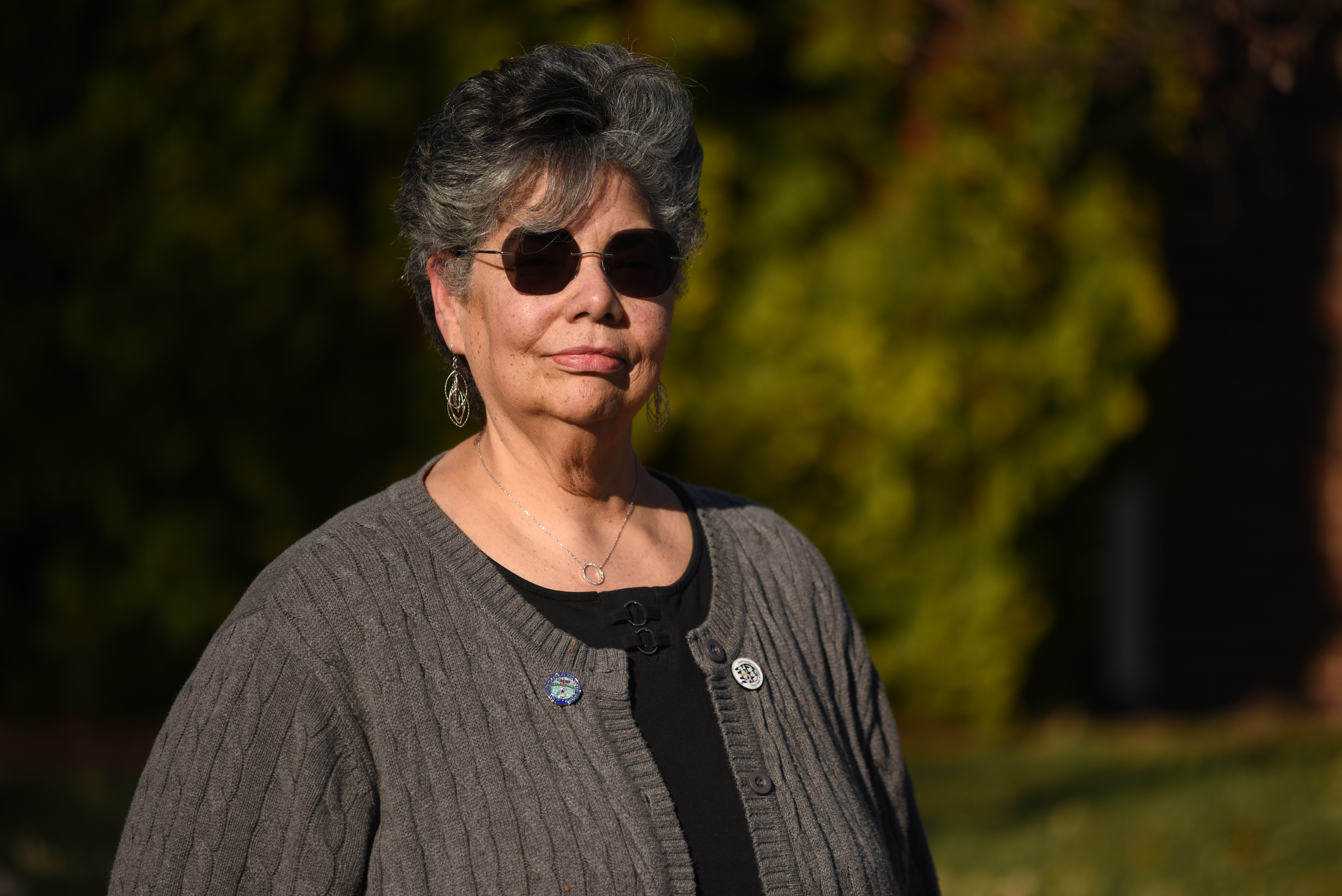 Tammy has short gray hair and sunglasses. She wears a gray sweater with two lapel pins.