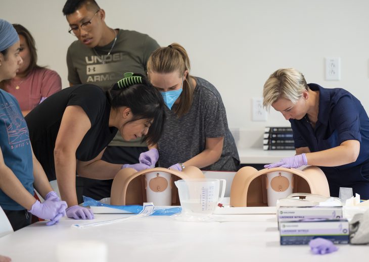 A student with medium black hair and a black t shirt, a student with long blond hair, a gray shirt and a mask and a student with short blond hair and blue shirt perform a procedure with mannequins on a table.