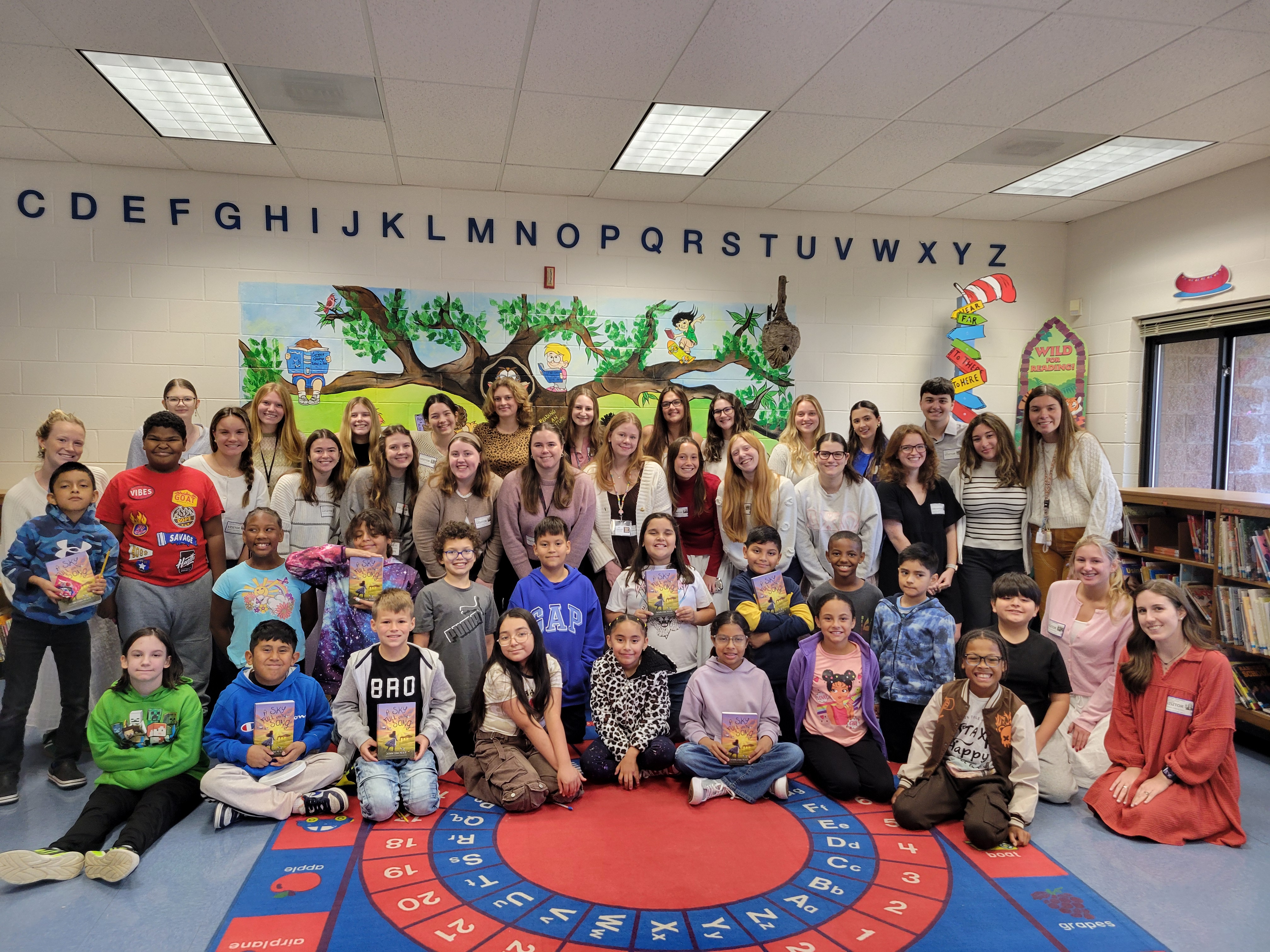 A large group of elementary students and adults pose together in a colorful classroom library, smiling for a group photo on an alphabet rug.