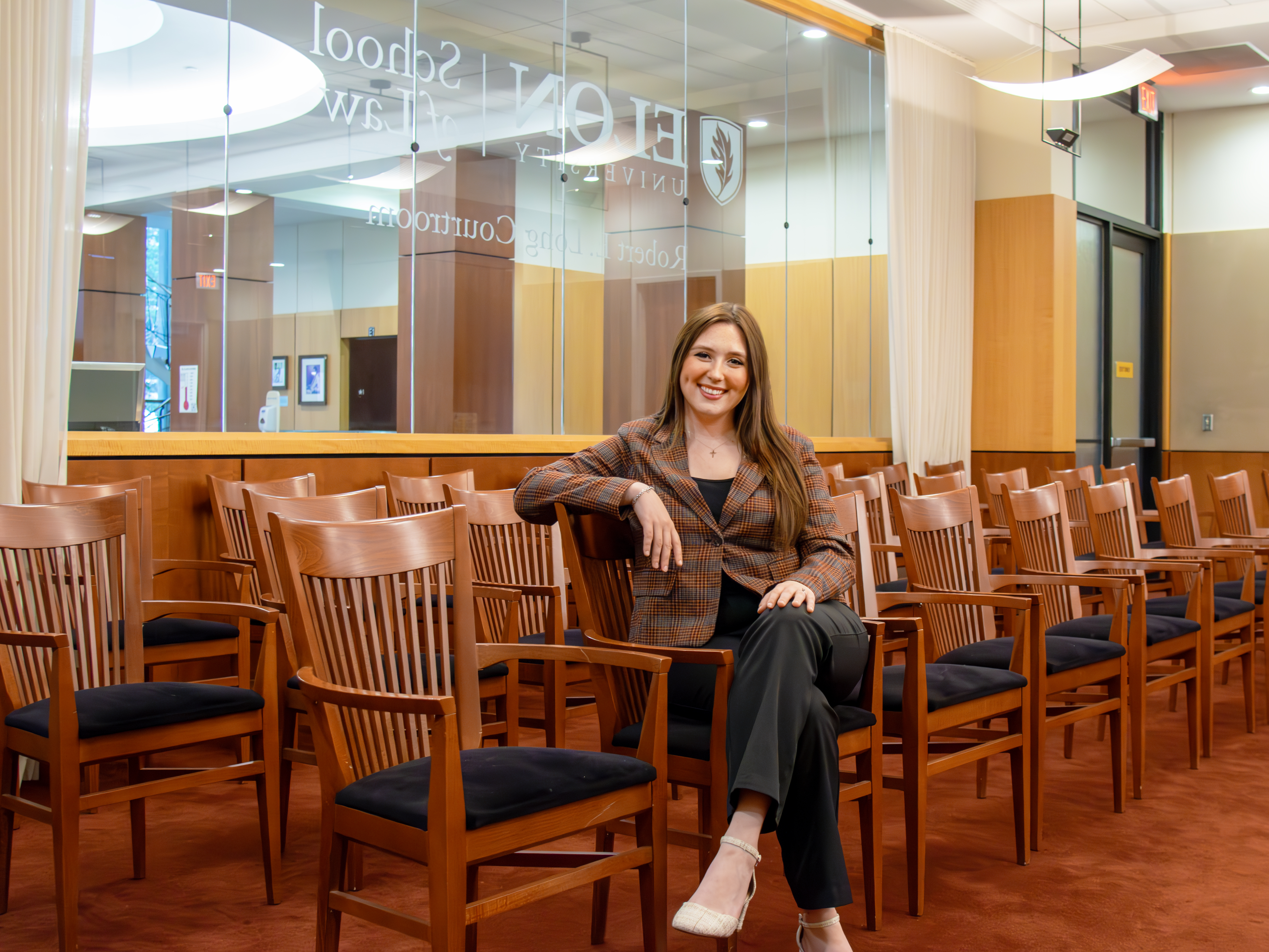 Rebecca “Becca” Bailey L’25 sits confidently in the Robert E. Long Courtroom at Elon University School of Law. She is smiling, dressed in a plaid blazer and black pants, seated among rows of wooden courtroom chairs with the courtroom’s glass wall and Elon Law signage visible behind her.