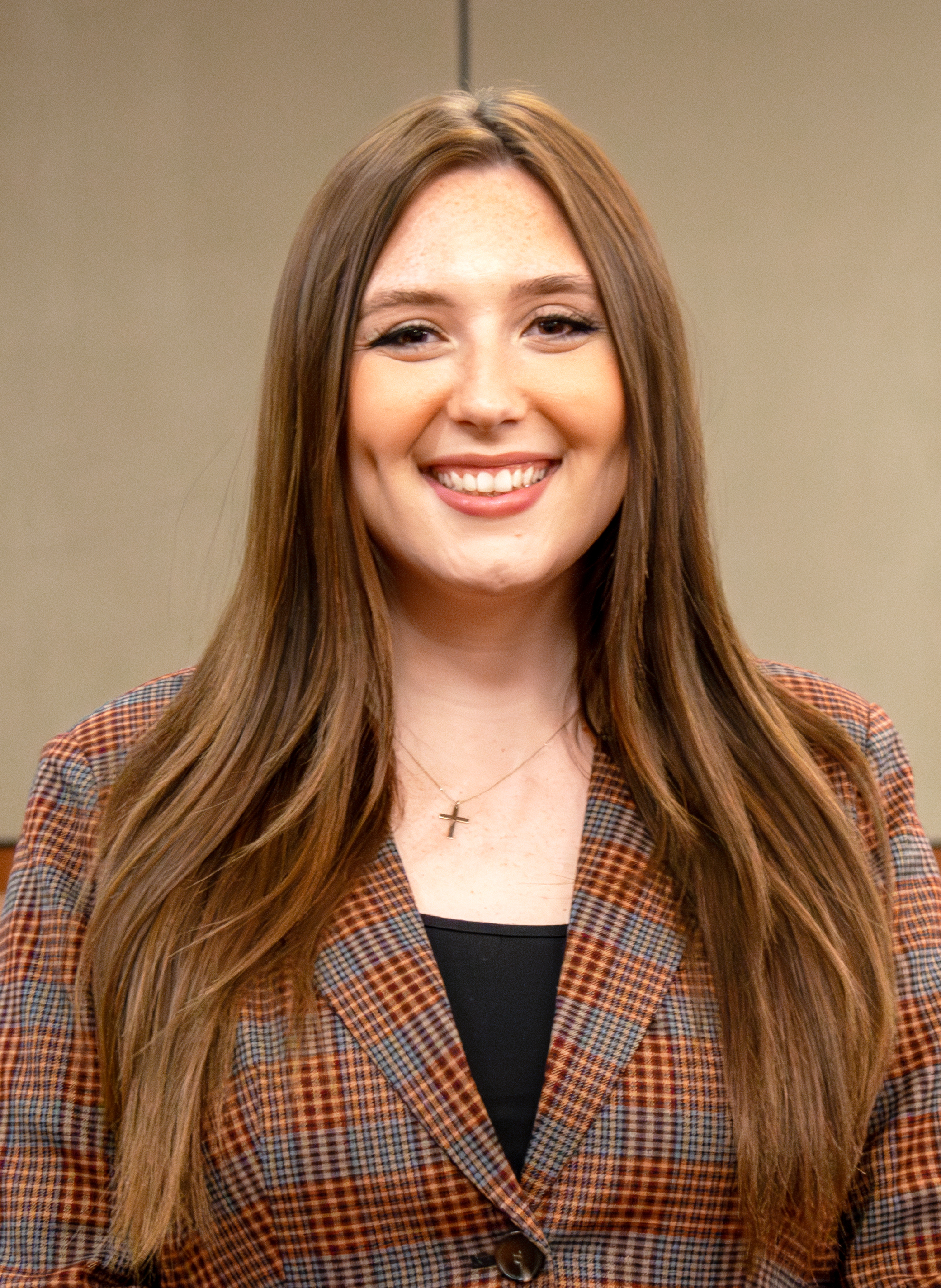 Rebecca “Becca” Bailey L’25 smiles for a headshot portrait. She is wearing a plaid blazer and a small cross necklace, with long brown hair parted at the center, standing against a neutral indoor background.