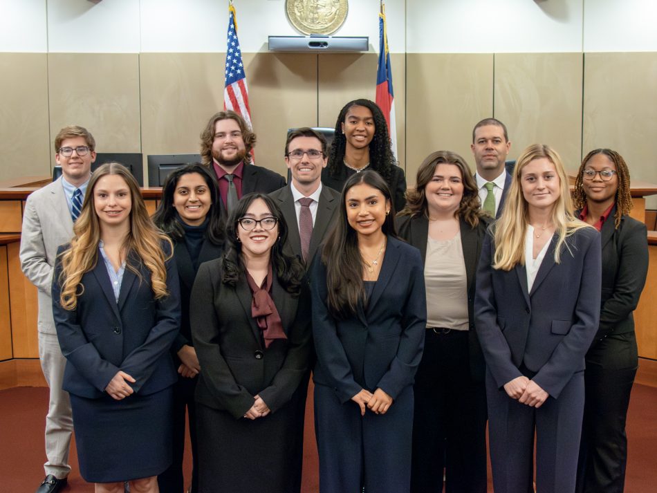 A group of students smiling in a courtroom, wearing professional dress.