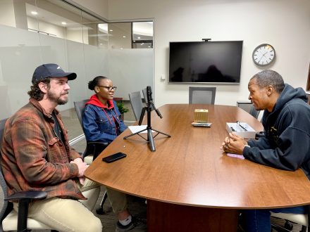 Two law students and a podcast host seated around a conference room table. The students are speaking into a microphone on the table.