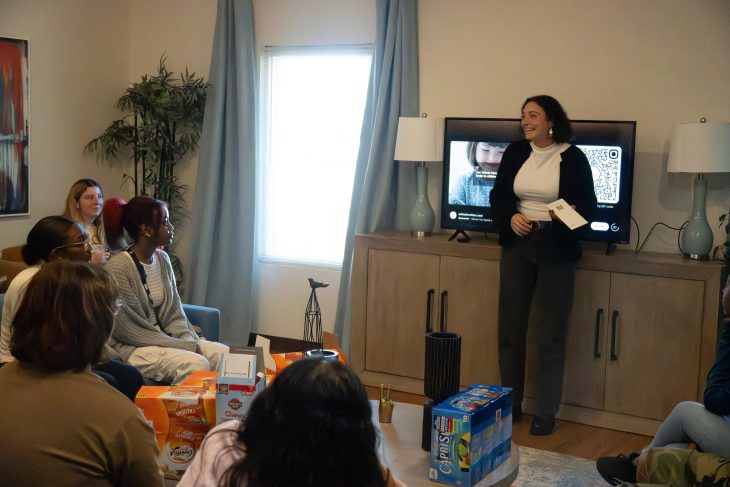 Junie Vargas ’21 stands at the front of a living-room-style space, smiling and speaking to a seated group of students. A TV behind them displays a slide with a QR code, and snacks are spread across a table as the group listens attentively