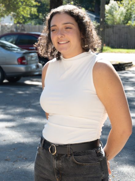 Junie Vargas ’21 stands outdoors in sunlight, smiling slightly while looking off to the side. They wear a sleeveless white top and dark jeans, with trees and parked cars softly blurred in the background.
