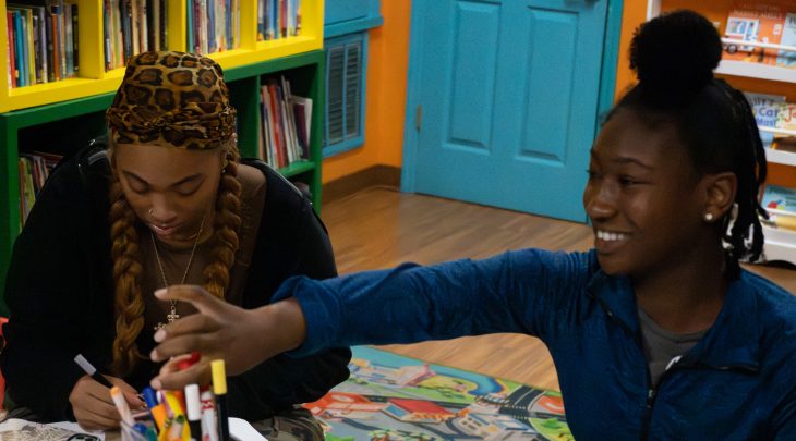 Two people sit at a table in a colorful reading room, working on an art activity with markers and paper. One person smiles while reaching for supplies as the other focuses on drawing, with bookshelves and children’s books in the background.