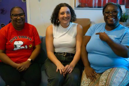 Angelia Stephens, shelter community family support coordinator, Junie Vargas ’21, and Lawana Crawford, lead resident advisor, sit side by side on a couch indoors, smiling toward the camera. Angelia wears a red graphic T-shirt, Junie wears a sleeveless white top, and Lawana wears a light blue T-shirt and gestures toward Junie.