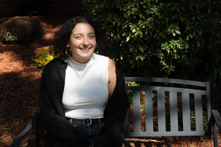 Junie Vargas ’21 sits smiling on a bench outdoors, wearing a sleeveless white top and black cardigan, with sunlight filtering through greenery behind her.