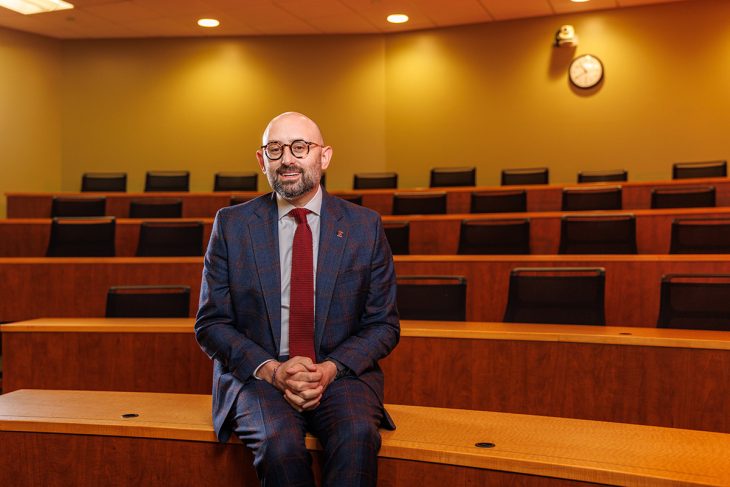 Zak Kramer, dean of Elon University School of Law, sits in a law school classroom, smiling toward the camera.