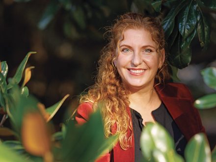 A faculty member smiles while standing among green foliage, sunlight filtering through the leaves.