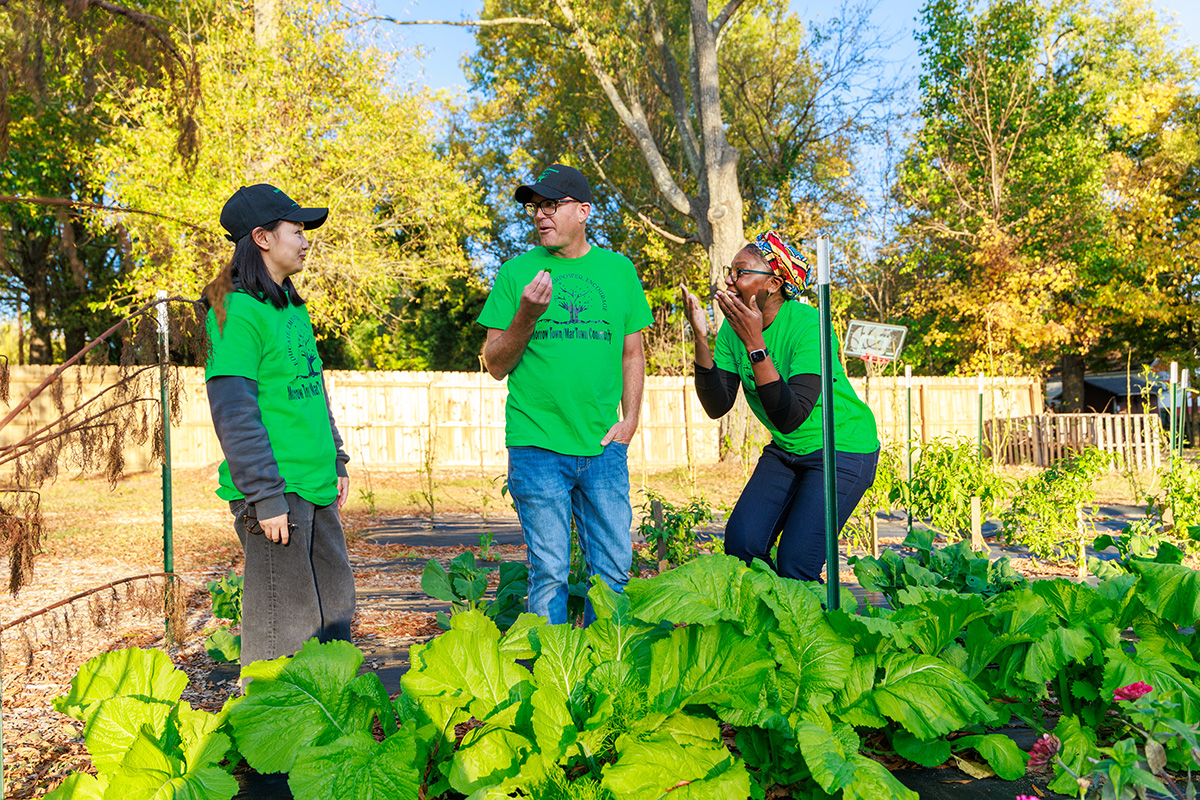 Three people talk and laugh while standing among leafy vegetables in a community garden.
