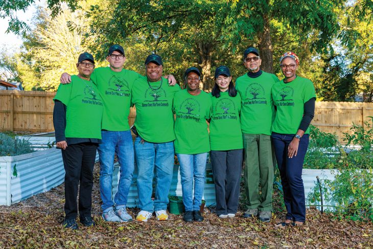 Volunteers wearing matching green shirts pose together in a community garden surrounded by raised beds.