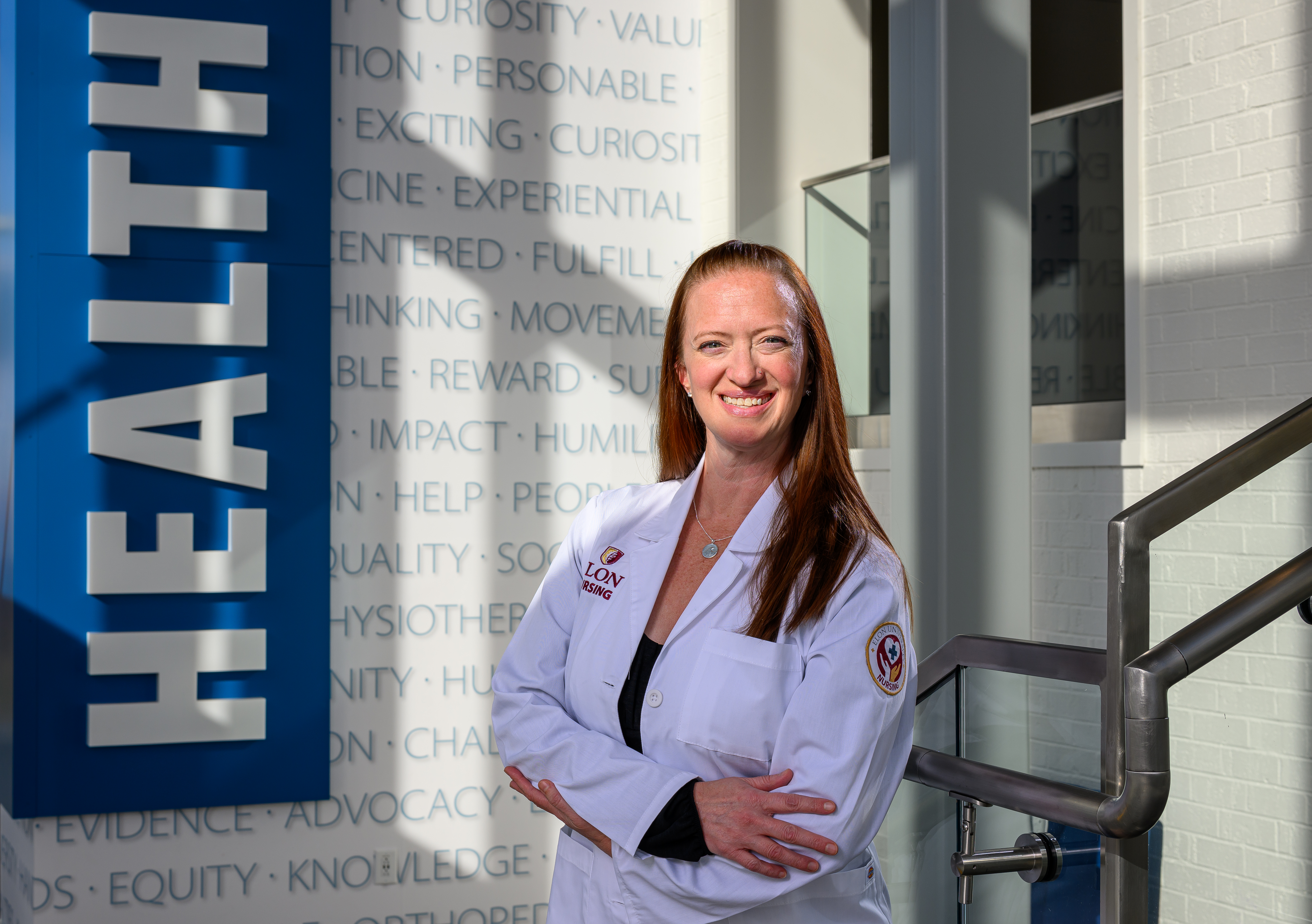 A woman in a white medical coat stands smiling with her arms crossed in a bright hallway next to a large vertical “HEALTH” sign. Sunlight falls across the wall behind her, which is covered in words related to healthcare and learning.
