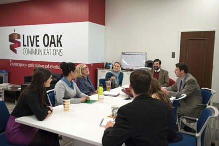Students and faculty sit around a table in the Live Oak Communications workspace, engaged in a group discussion.