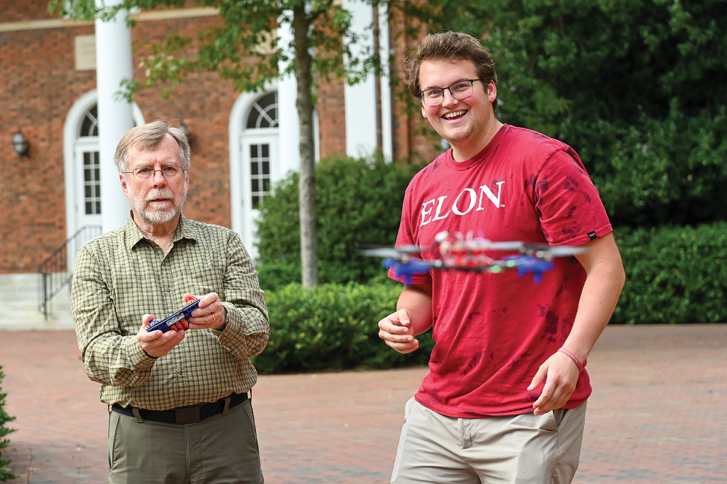 A student smiles as a faculty member operates a drone during an outdoor learning activity on campus.