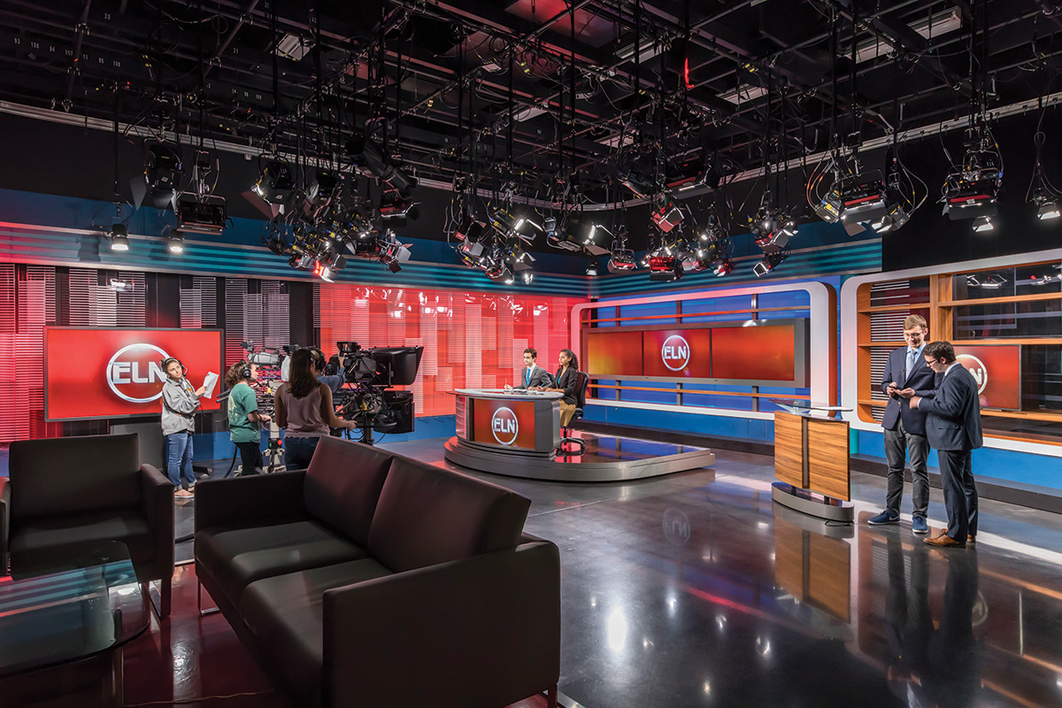 Students prepare for a broadcast inside a professional television studio with cameras, lights and an anchor desk.