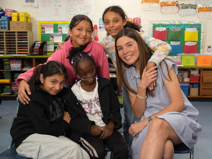 A teacher sits beside four students who lean in close, all smiling together in a classroom setting.