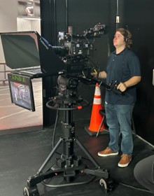Alt text: A person operates a professional studio camera setup with multiple monitors, standing beside an orange traffic cone on a production set.
