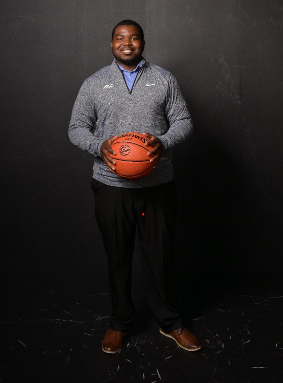 Alt text: A person stands smiling against a dark background while holding a basketball in both hands.
