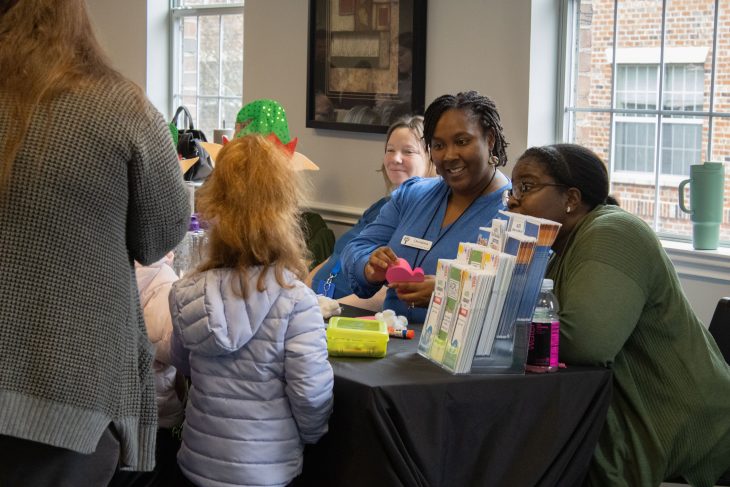 Alt text: Adults sit behind a table at a community event, smiling and interacting with children as they share information and hands-on activities inside a bright room with large windows.