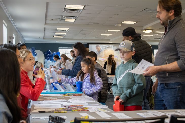 Children and adults gather around a long table at an indoor event, browsing books and materials while volunteers assist them, with balloons and activity stations visible in the background.