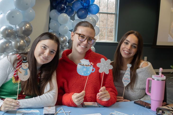 Three people sit together at a table smiling and holding winter-themed photo props, with blue and silver balloons decorating the space behind them.