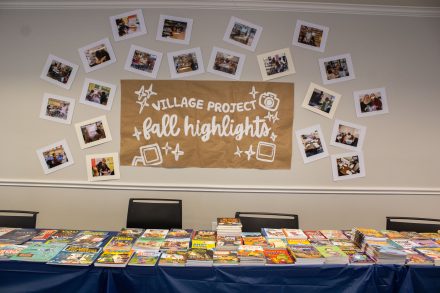 Alt text: A display table is covered with stacks of books, with photos arranged on the wall above around a sign reading “Village Project Fall Highlights.”