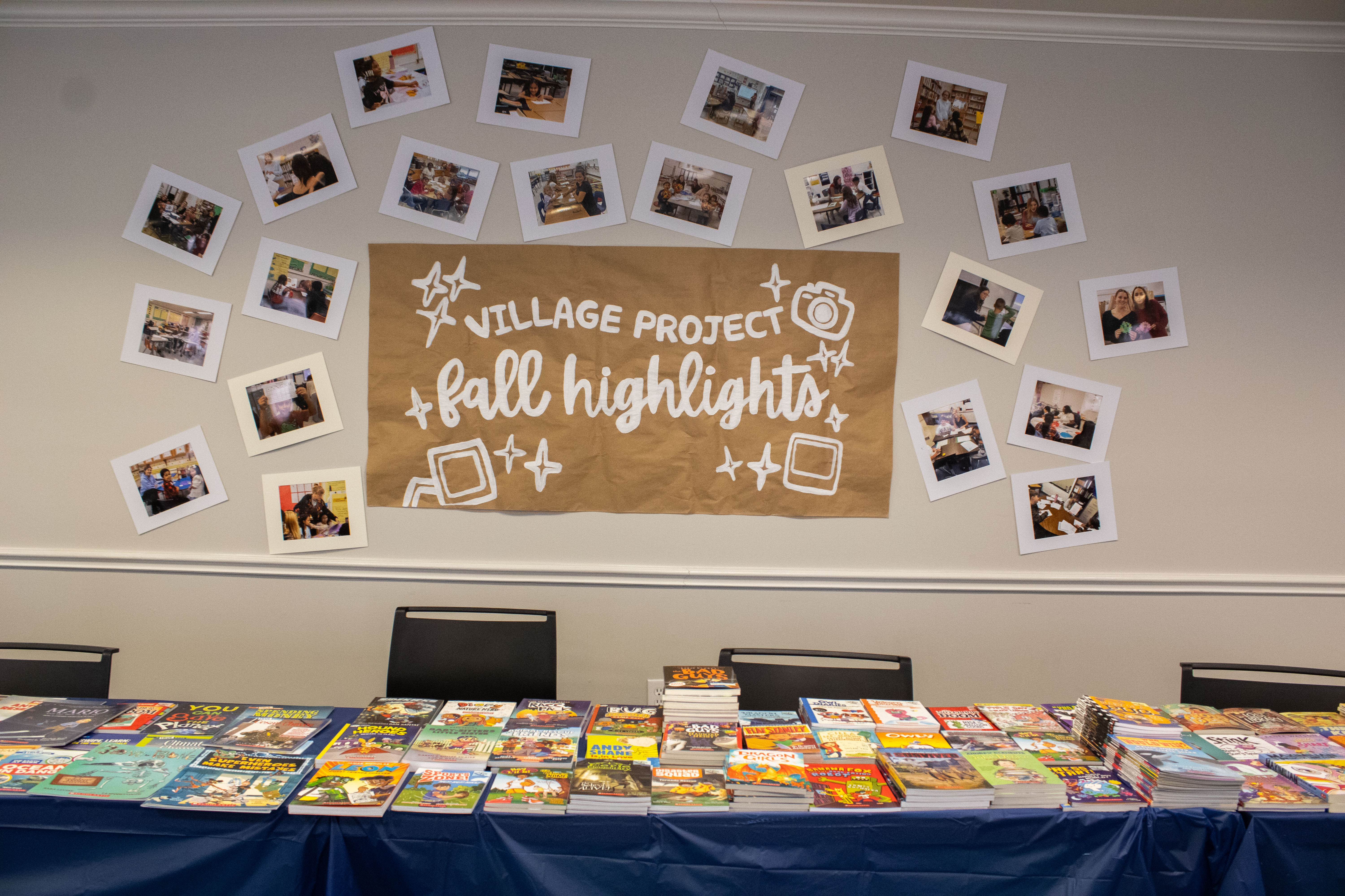 Alt text: A display table is covered with stacks of books, with photos arranged on the wall above around a sign reading “Village Project Fall Highlights.”