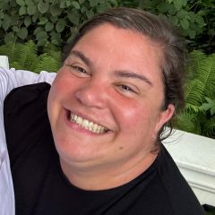 A young woman wearing a black T-shirt with her hair in a ponytail smiles up at the camera in a selfie.