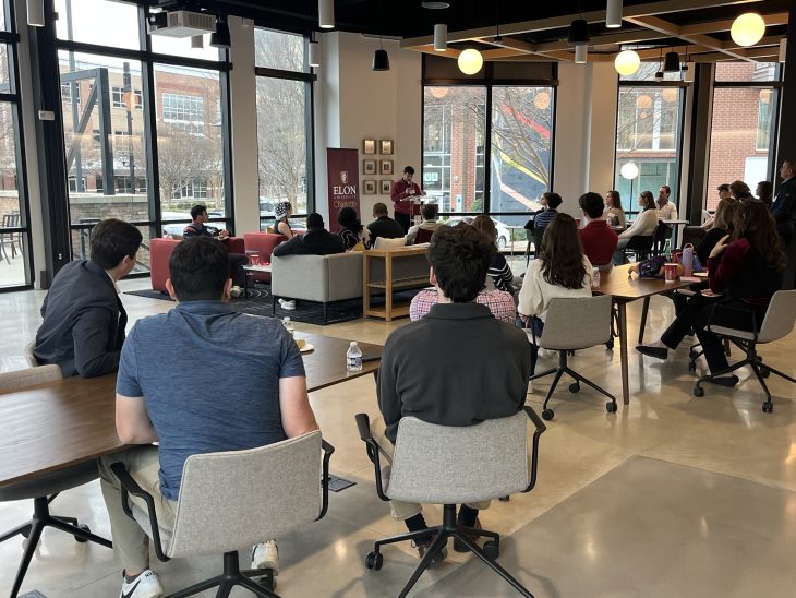 People sit at tables and chairs in a modern, open room listening to a speaker at the front, with large windows and campus buildings visible outside.