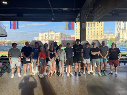 A group of people stand in a line under a covered outdoor structure at a sports venue, with a city skyline and flags visible in the background.