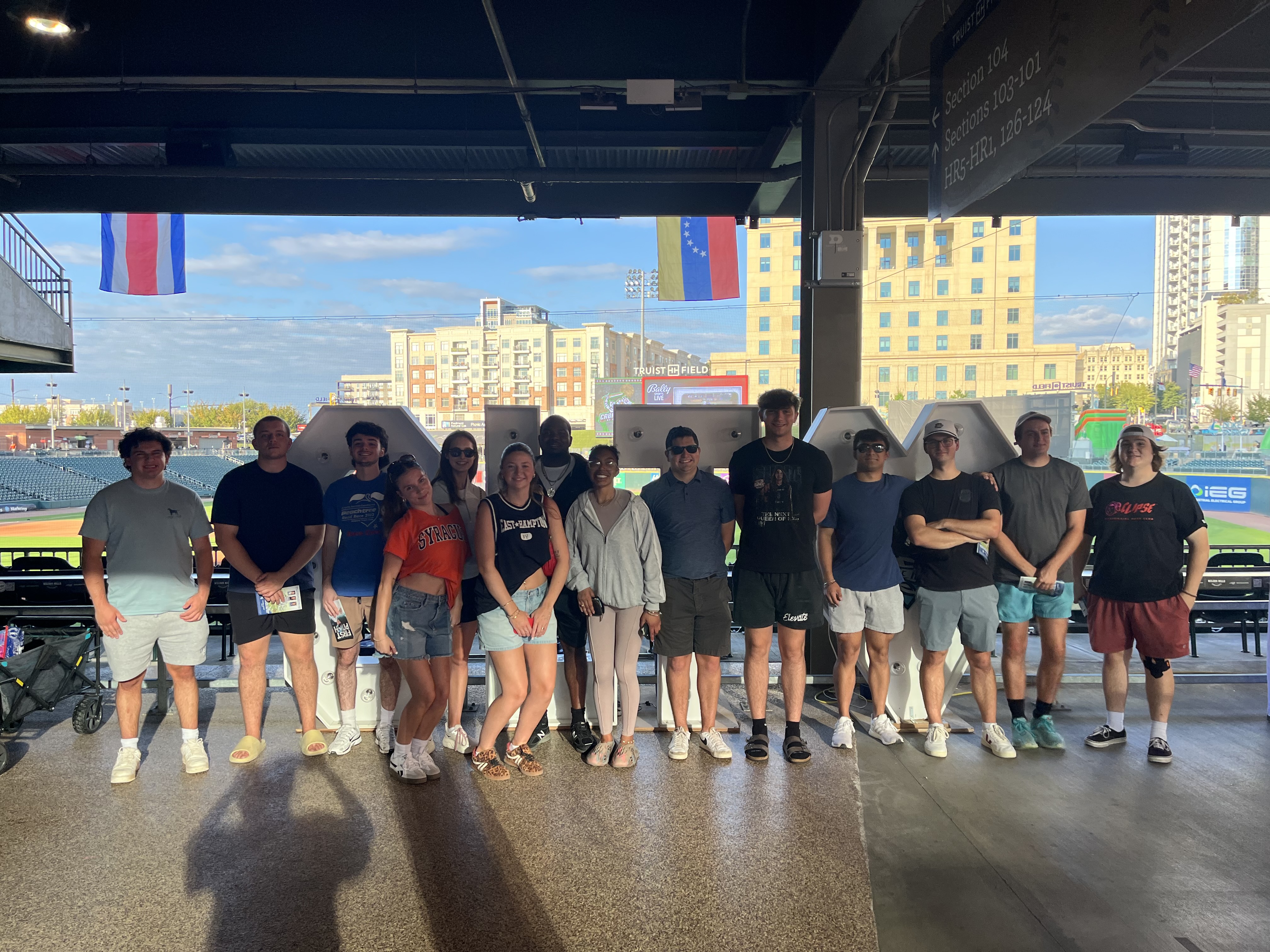 A group of people stand in a line under a covered outdoor structure at a sports venue, with a city skyline and flags visible in the background.
